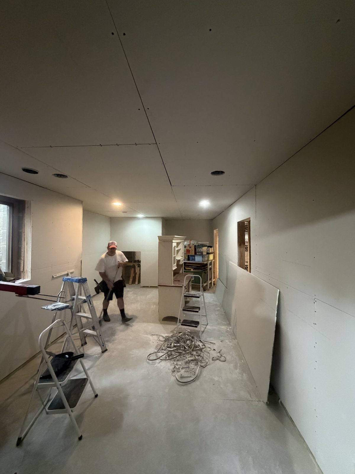 Long basement room fully boarded in drywall with a worker sweeping the concrete floor