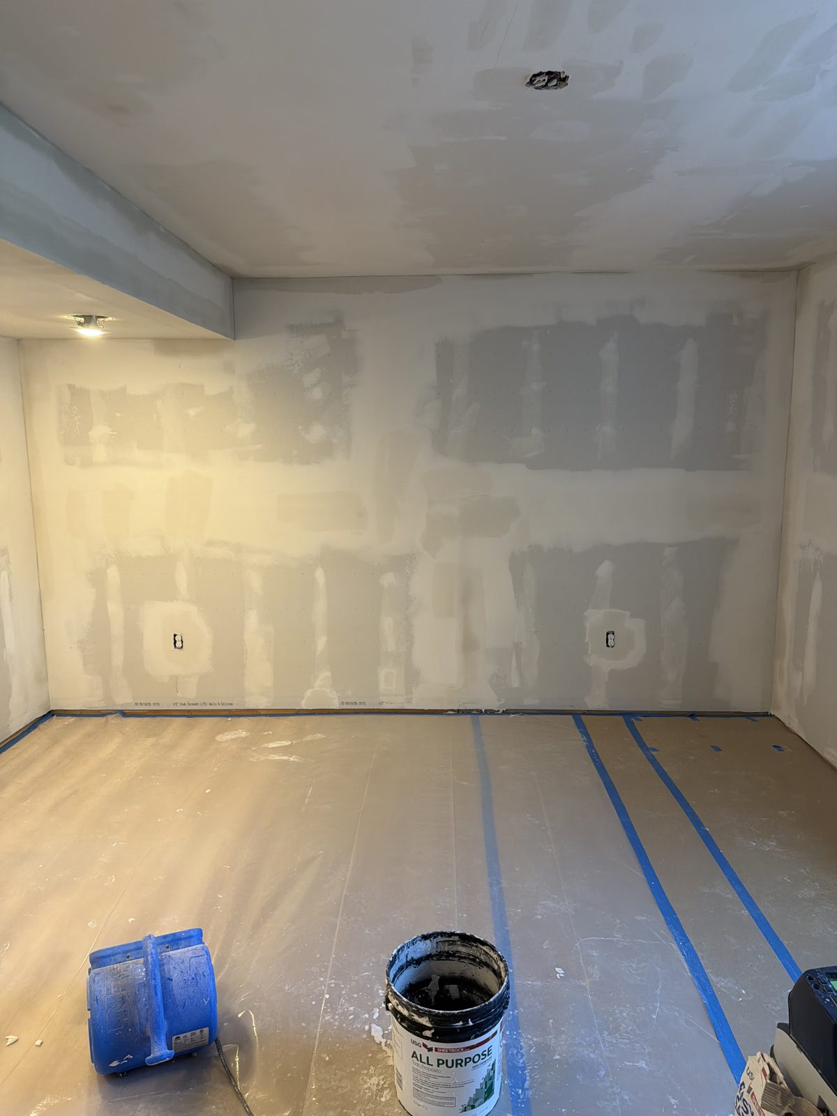 Basement wall with all drywall seams and screw heads coated in joint compound, floor covered in plastic