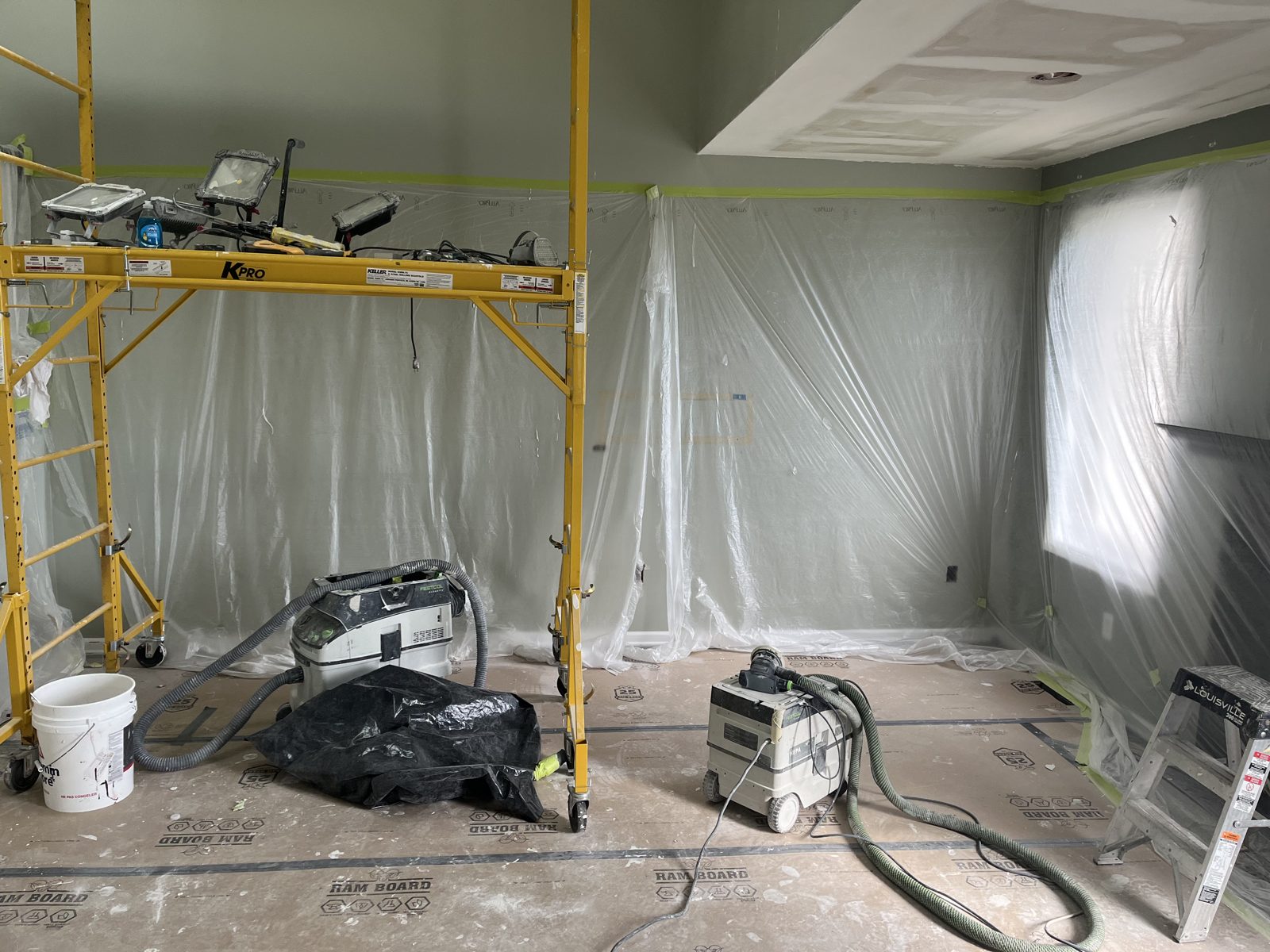 Living room fully masked with plastic sheeting and floor protection, scaffolding and dust extractors staged for drywall work
