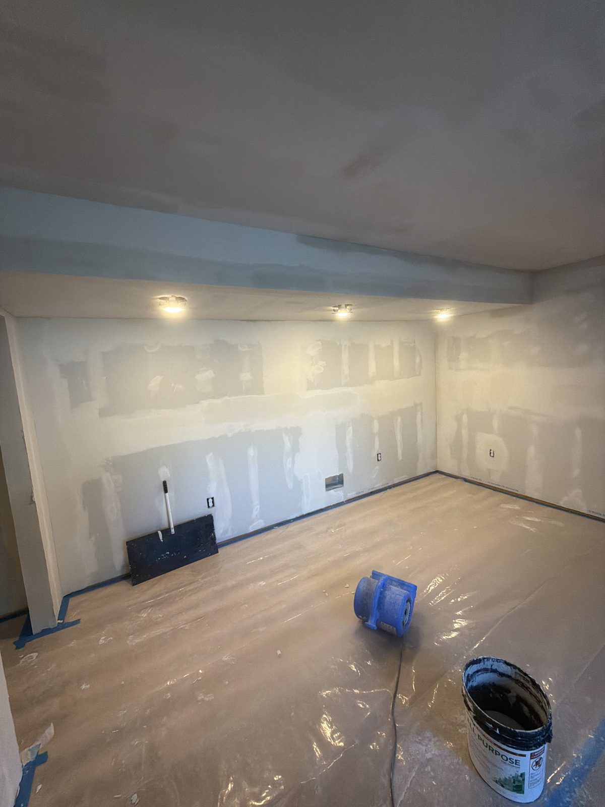 Basement room with taped drywall corners and a coated soffit, protected floor and joint compound bucket